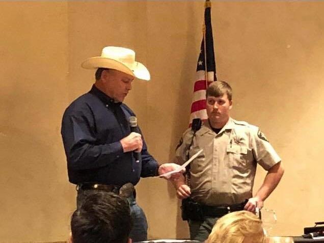 Sheriff Tanner standing with a microphone reading from a sheet of paper. Deputy Ryan Gantt stands to the side